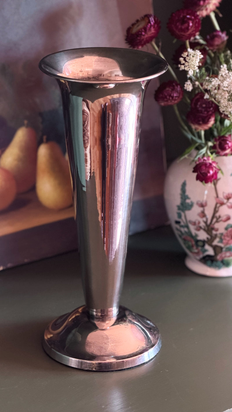 A polished silver-toned metal flared vase or goblet with a circular base, shown on a tabletop with a blurred floral arrangement and fruit in the background.