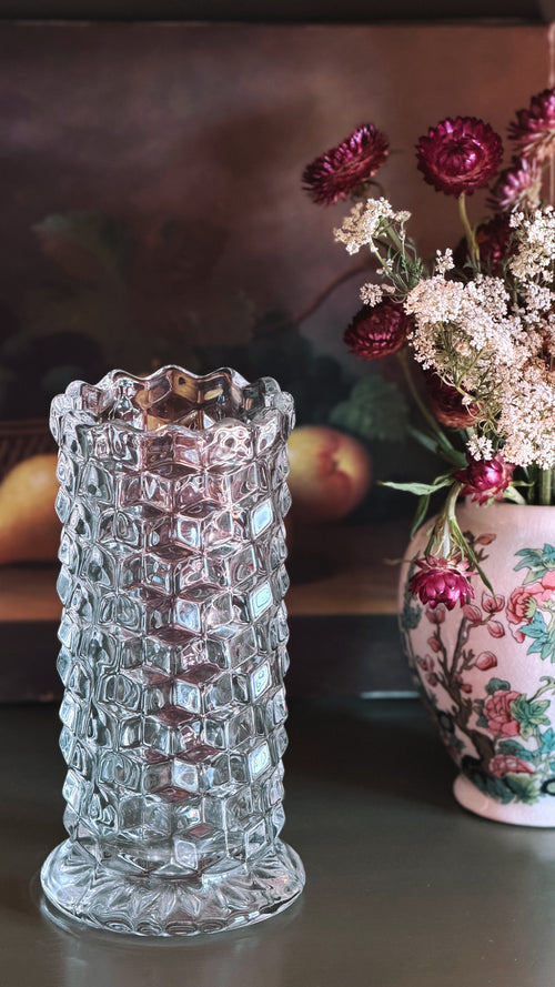 Fostoria American clear glass straight vase, 8 inches tall with a diamond-patterned body and flared rim, shown on a tabletop with dried flowers in a floral pot blurred in the background.