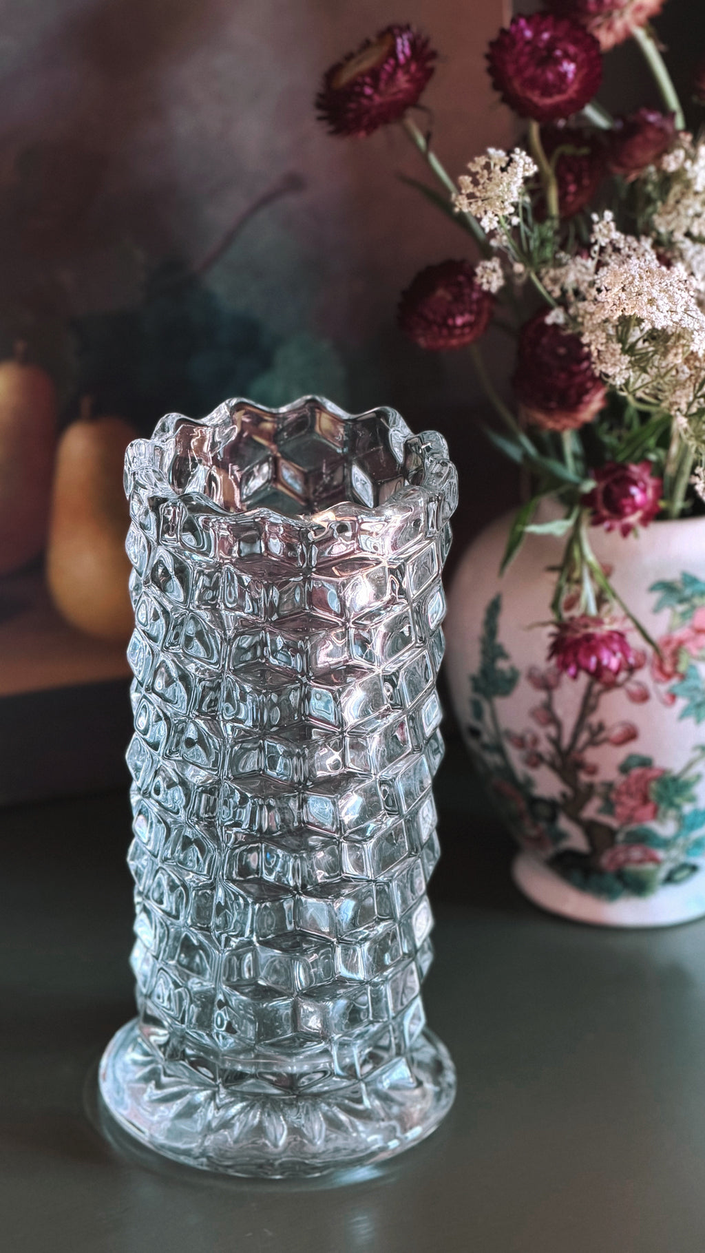 A clear textured glass vase with a diamond-patterned body and flared base, shown on a tabletop with dried flowers in the background.