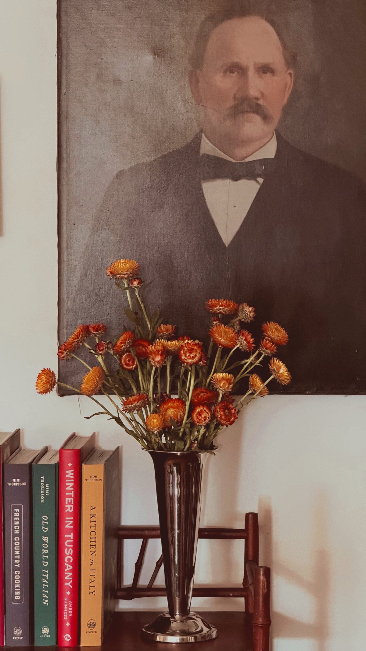 A vintage-style portrait painting of a man with a mustache hangs on a wall above a table holding a tall dark vase filled with orange flowers, next to a row of books.
