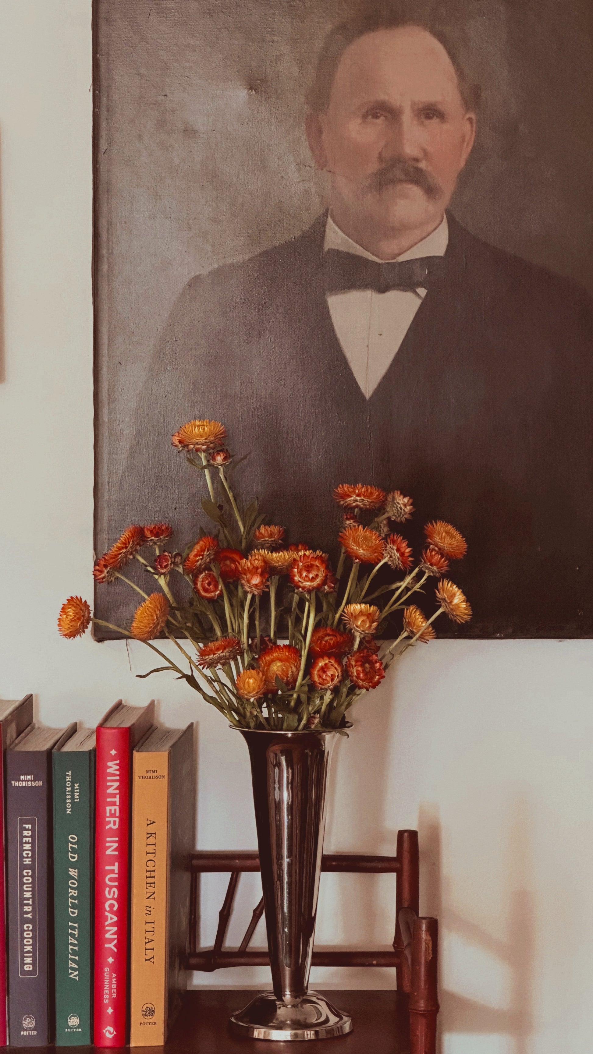 A vintage-style portrait painting of a man with a mustache hangs on a wall above a table holding a tall dark vase filled with orange flowers, next to a row of books.