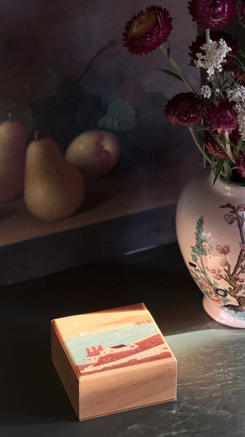 A square wooden keepsake box with a colorful landscape illustration on the lid sits on a dark tabletop. In the softly lit background, a ceramic vase with flowers and pears are visible.