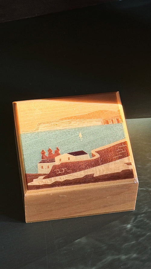 A square wooden box with a hand-painted scenic coastal landscape design on the lid, resting on a dark reflective surface.