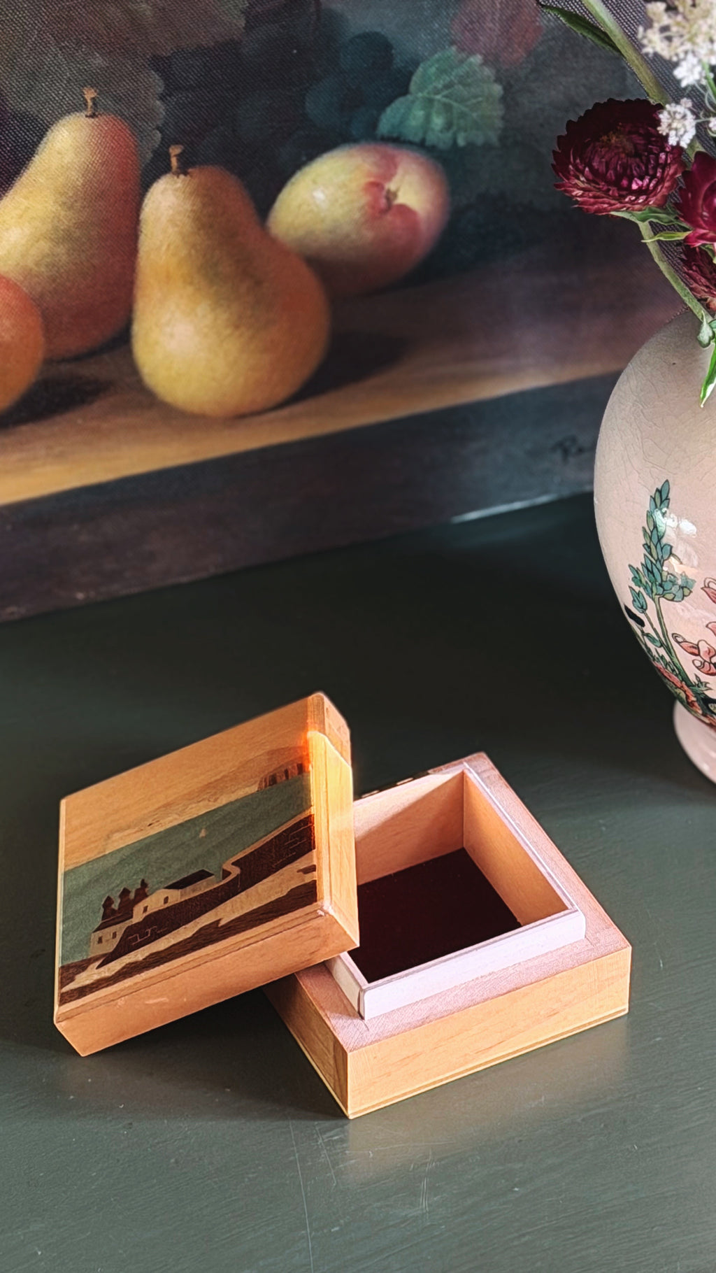 A square wooden keepsake box with a hinged lid, shown open on a dark tabletop. The lid features an illustrated scene of a seaside village, while the inside of the box is lined with pink velvet.