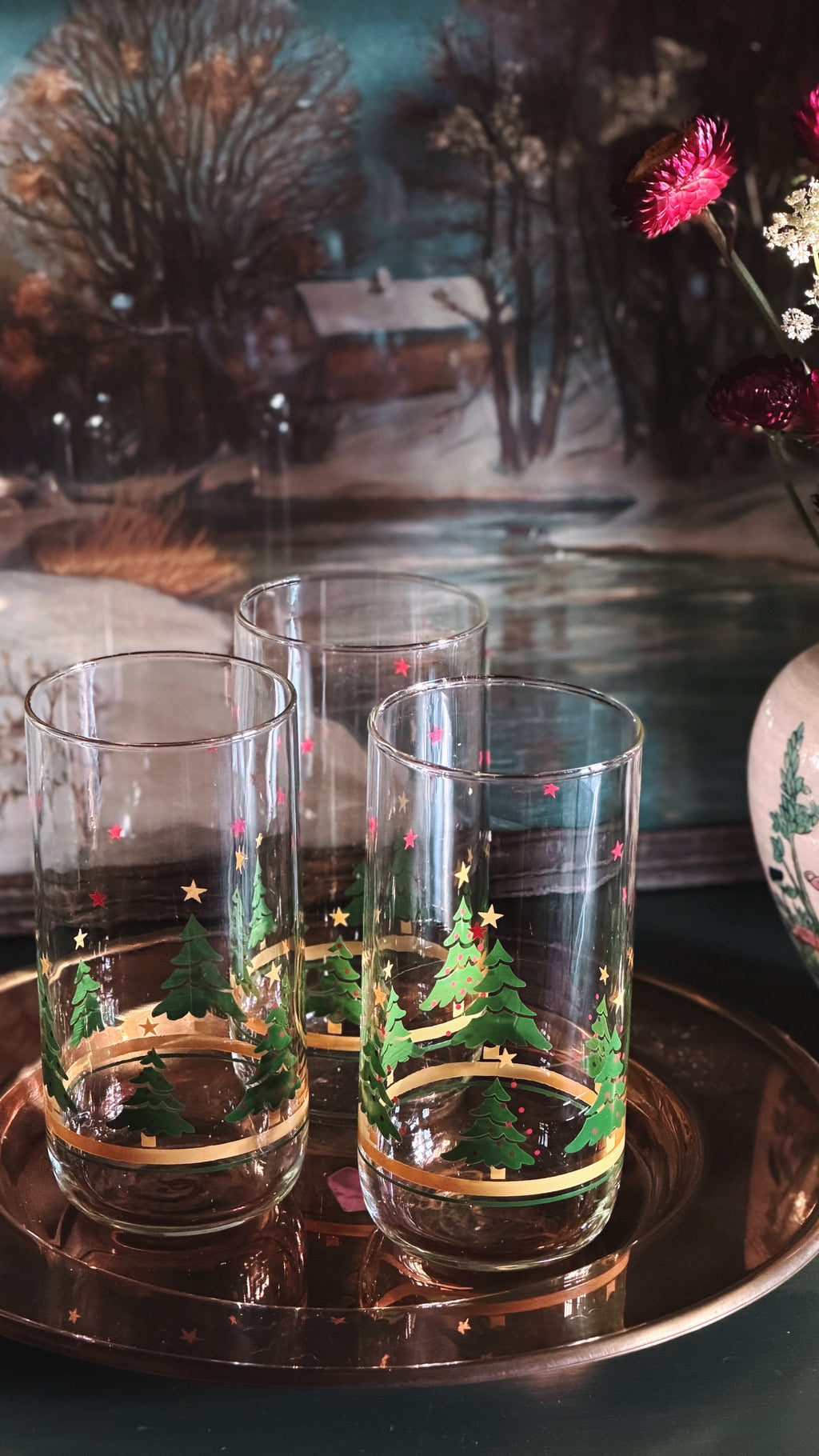 Vintage Christmas Tree Gold Band Glass Tumblers with hand-painted green Christmas trees, gold stars, and gold bands, arranged on a copper tray against a rustic, blurred background.
