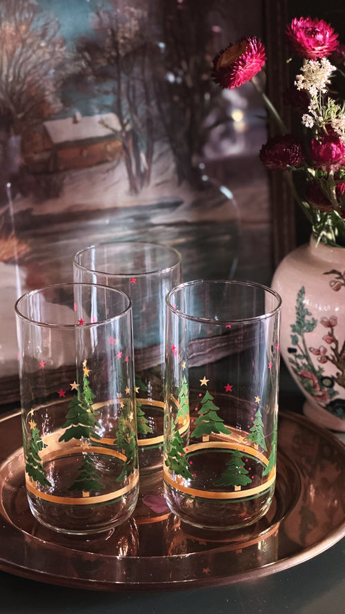 Vintage Christmas Tree Gold Band Glass Tumblers with green Christmas tree and gold circular band designs, arranged on a copper tray with a blurred floral and scenic background.