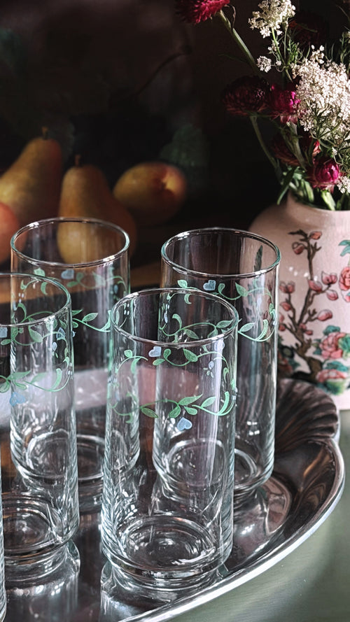 Vintage Corelle Country Cottage 16 oz Tumbler with delicate green floral vine patterns, arranged on a silver tray with a dark, rustic background featuring pears and a floral vase.