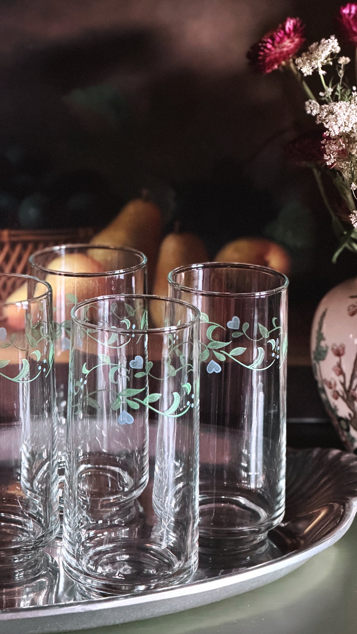 Vintage Corelle Country Cottage 16 oz Tumblers with delicate green and blue floral vine patterns, arranged on a silver tray with a warm, dark, rustic background.