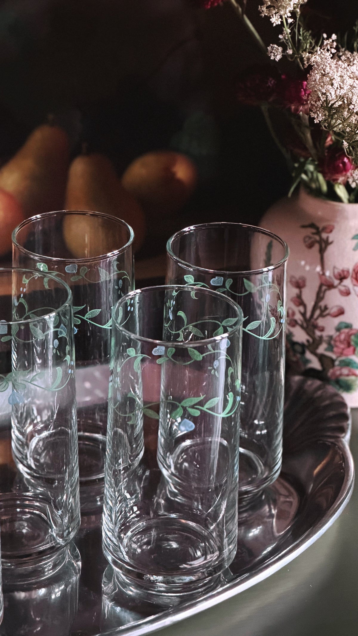 Four clear glass tumblers with delicate light green floral vine patterns, arranged on a reflective silver tray with a dark, softly blurred background featuring pears and a vase of flowers.