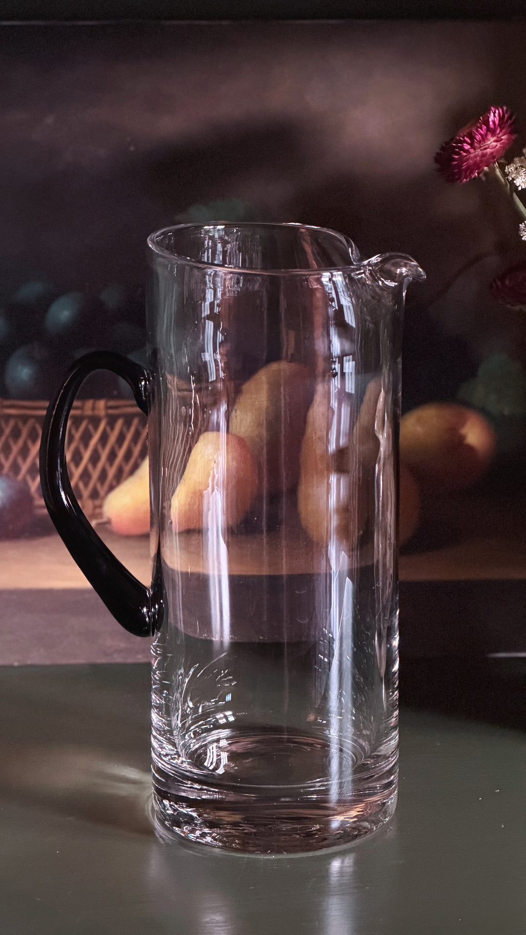 A clear glass pitcher with a black handle and spout, shown on a reflective surface with a softly blurred dark background and fruit props.
