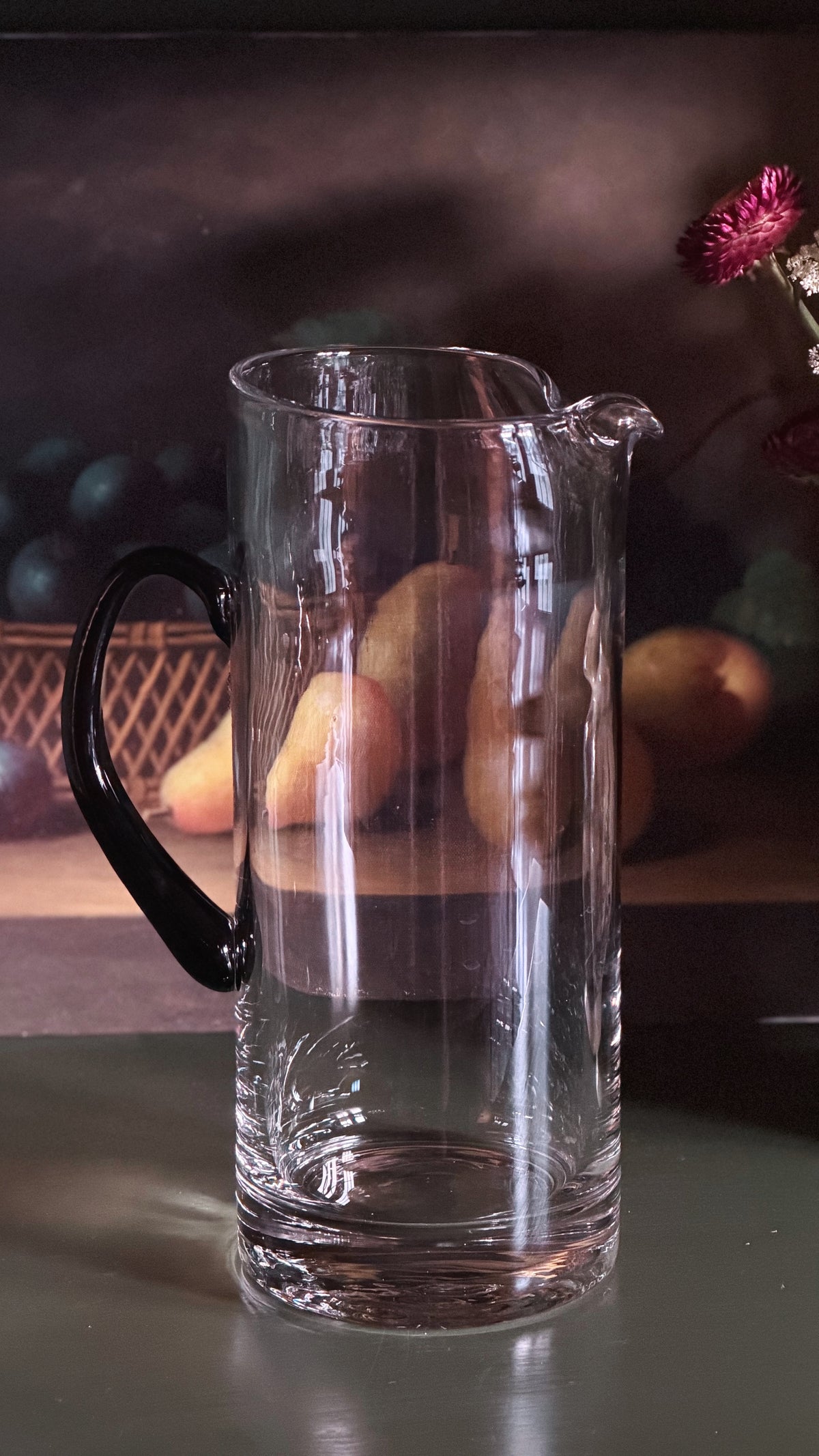 A clear glass pitcher with a black handle and spout, shown on a reflective surface with a softly blurred dark background and fruit props.