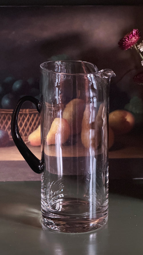 A clear glass pitcher with a black handle and spout, shown on a reflective surface with a softly blurred dark background and fruit props.