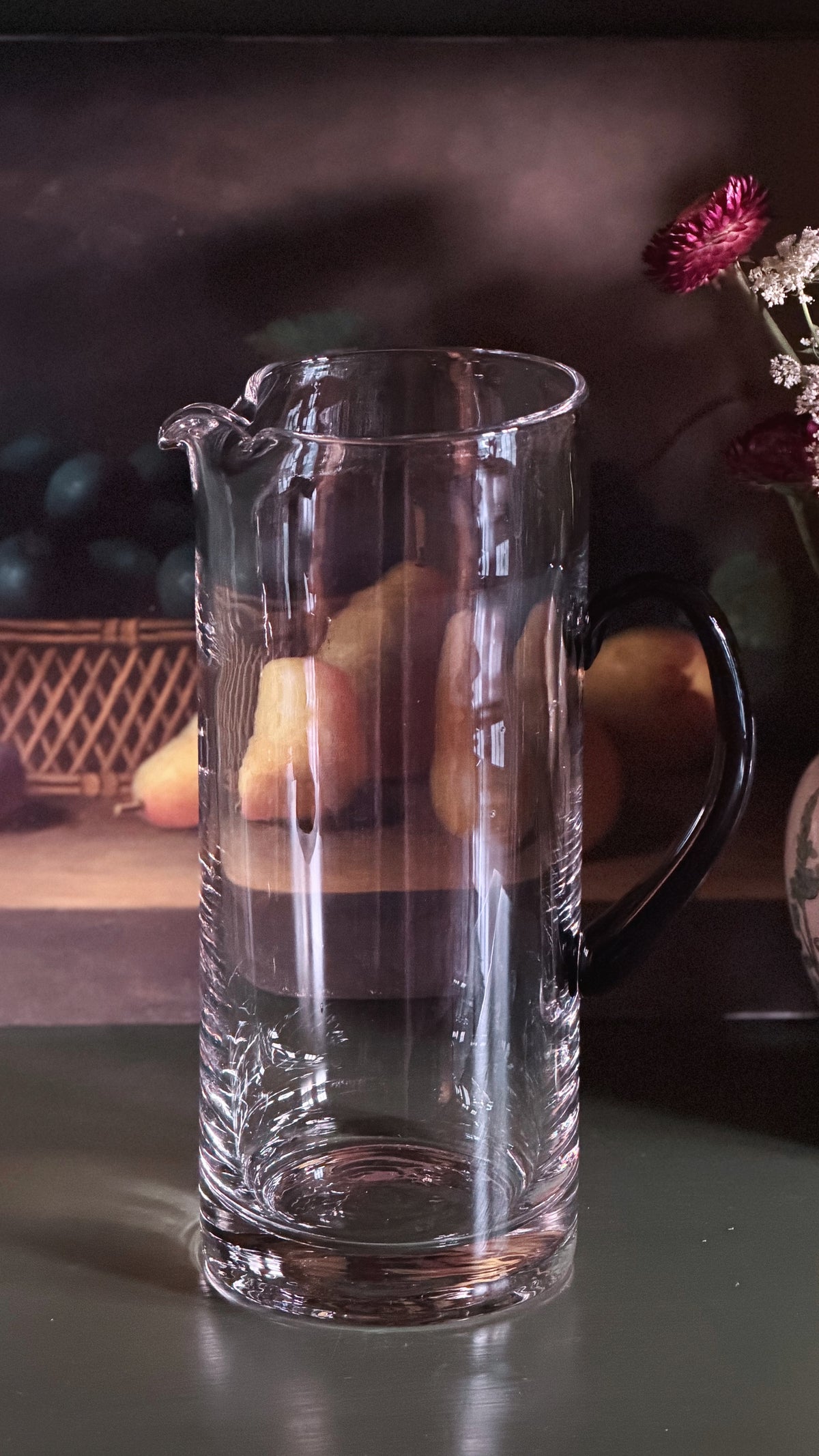 A clear glass pitcher with a black handle and spout, shown on a dark tabletop with softly blurred fruit and flowers in the background.