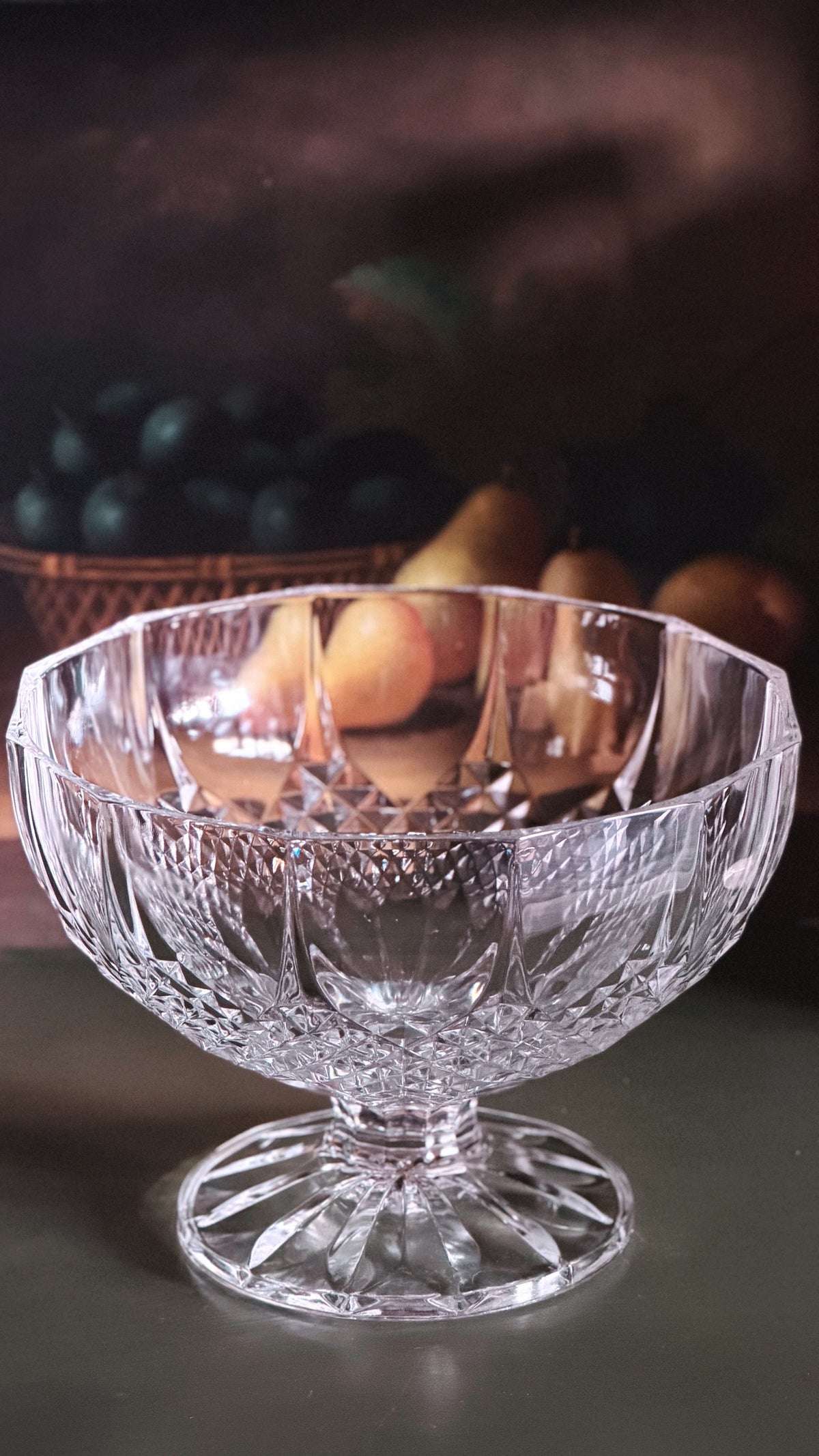 A clear cut-crystal pedestal bowl with a flared base, shown on a dark tabletop with fruit blurred in the background.