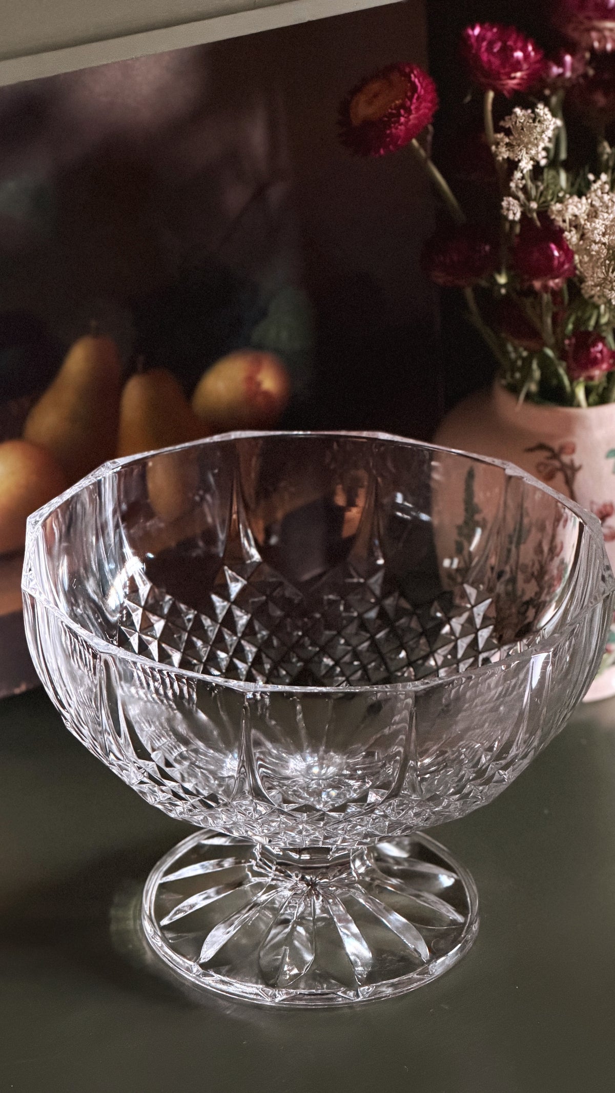 A clear cut-crystal pedestal bowl with a diamond-patterned body and flared base, photographed on a tabletop with flowers and fruit blurred in the background.