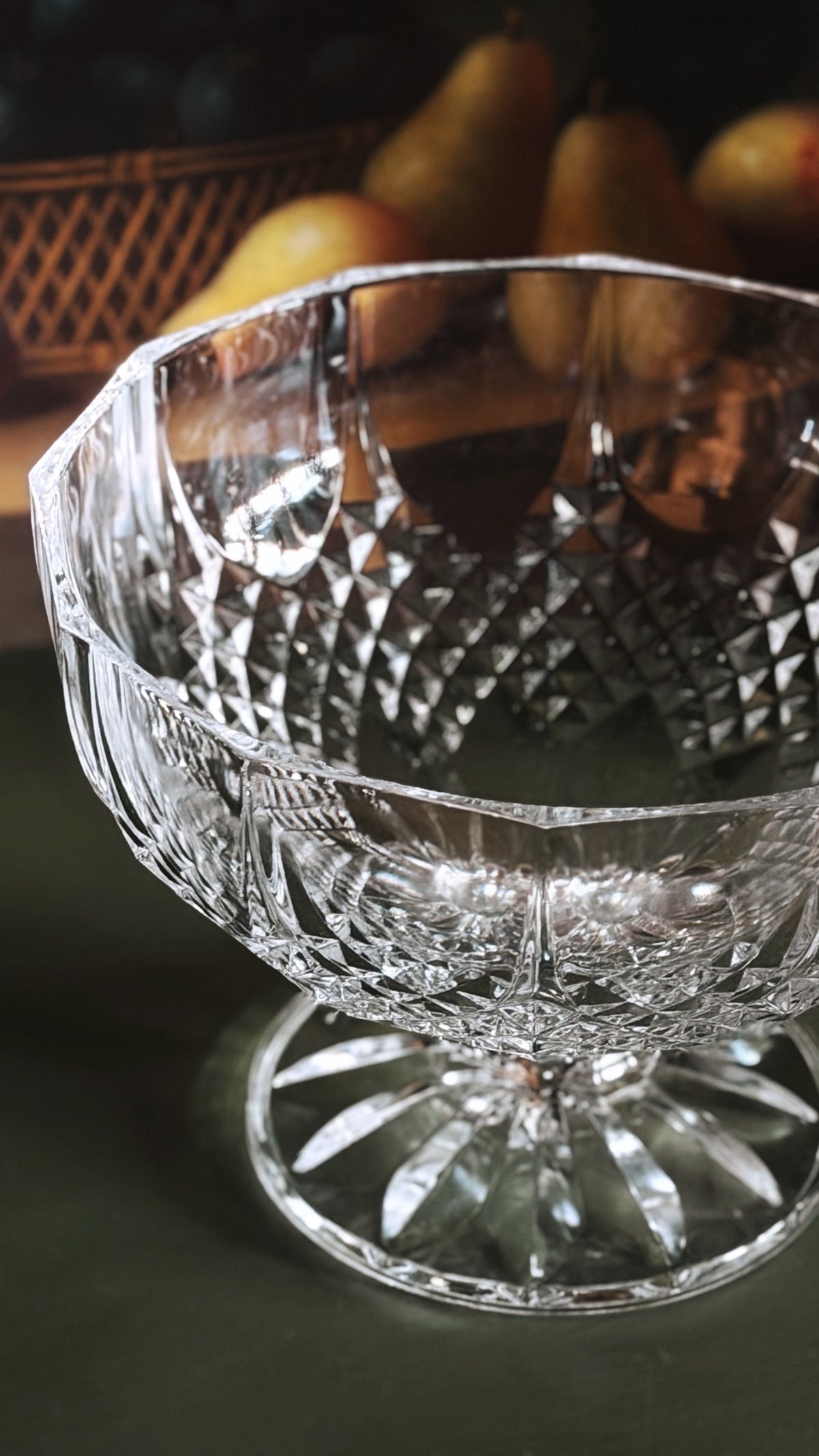 A clear cut-crystal pedestal bowl with a faceted base, shown in a close-up studio-style shot with softly blurred fruit in the background.