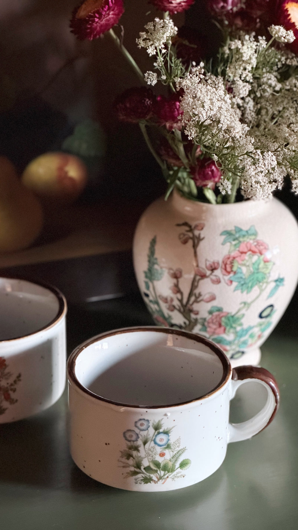A white speckled ceramic mug with a floral print and brown rim in the foreground, with two matching mugs and a floral-patterned vase filled with dried flowers on a dark tabletop.