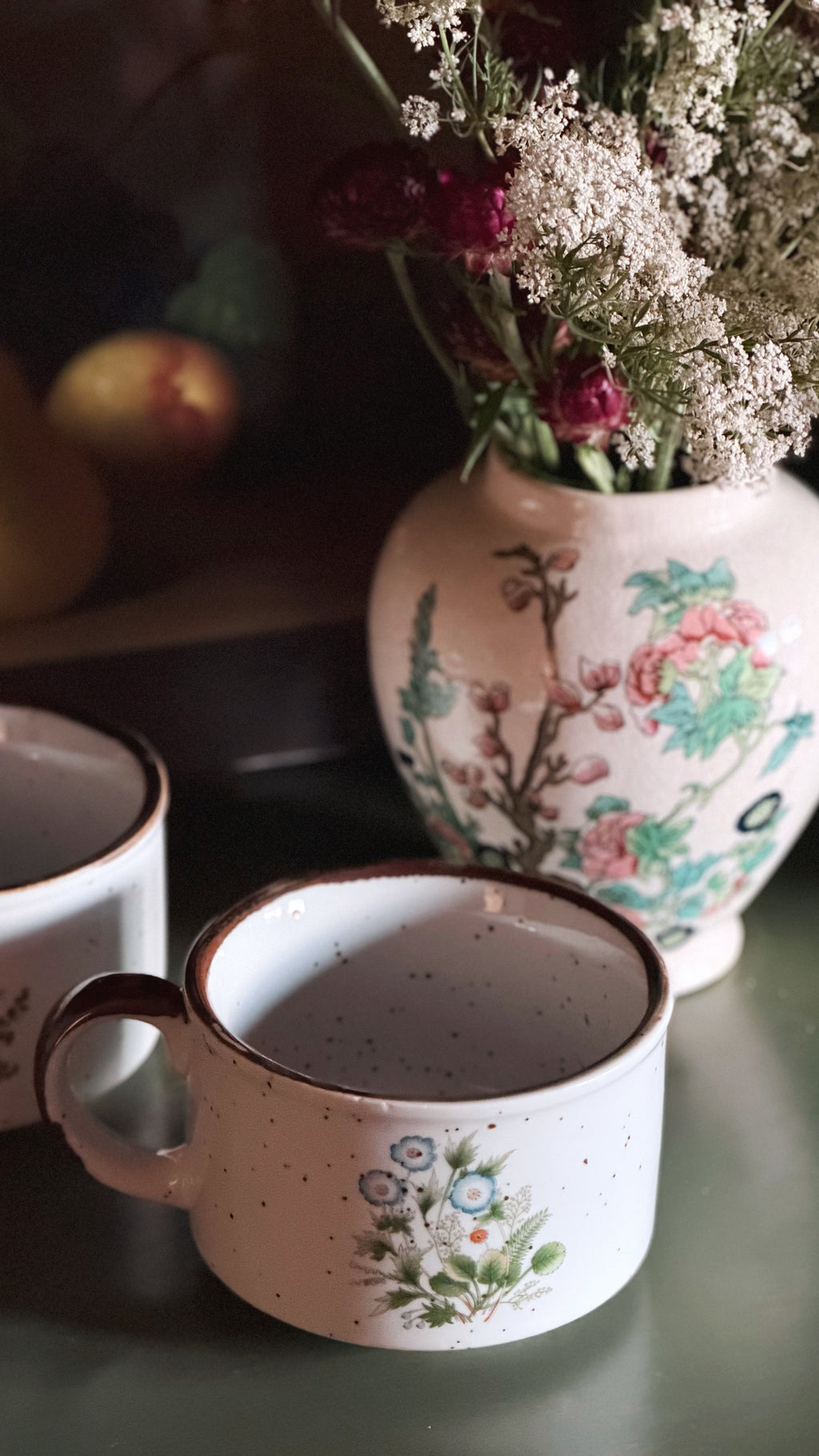A close-up of a speckled ceramic mug with a floral print, placed on a dark green surface next to another mug, with a larger floral vase and dried flowers in the background.
