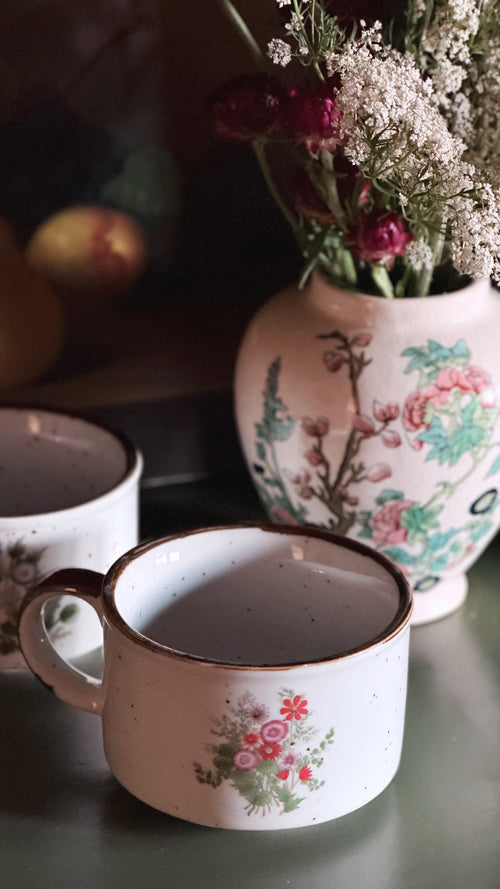 A white ceramic mug with a floral print and brown rim in the foreground, with two matching mugs and a floral vase in a softly lit setting.