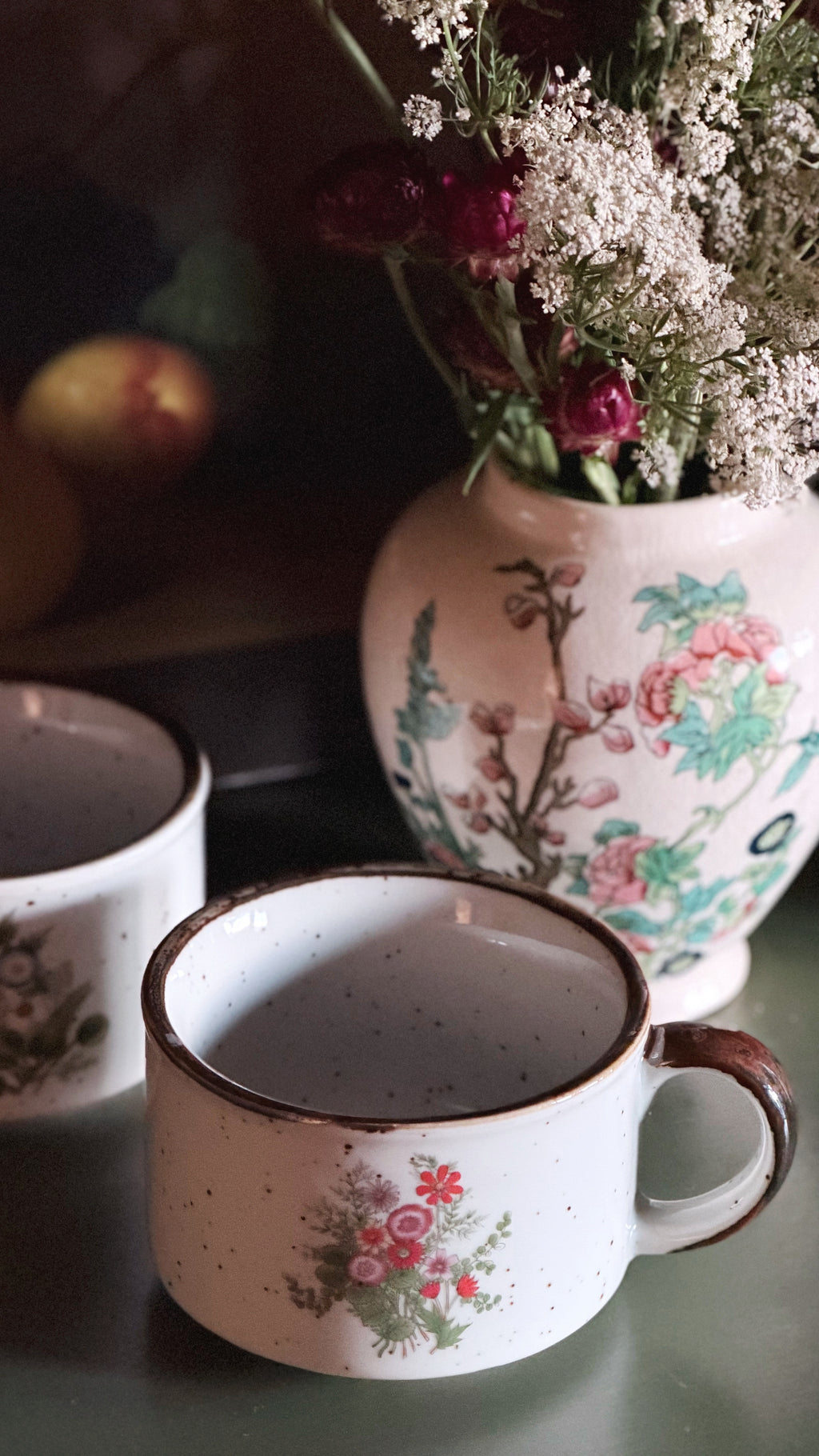 A close-up of a speckled white ceramic mug with a floral print and brown rim, set on a dark surface next to another matching mug and a floral vase in the background.