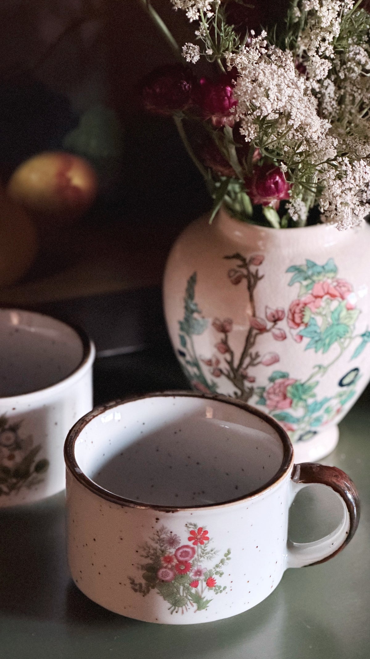 A close-up of a speckled white ceramic mug with a floral print and brown rim, set on a dark surface next to another matching mug and a floral vase in the background.