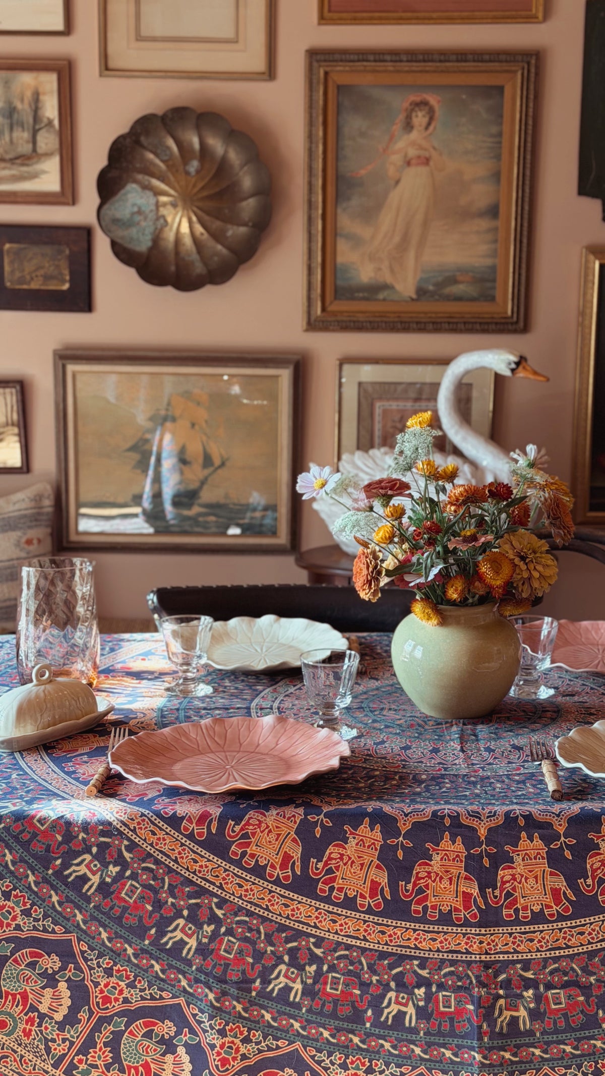 A styled dining table in a warm room setting, featuring a navy blue tablecloth with red and gold elephant motifs, pink plates, a floral centerpiece in a green vase, and a white swan figurine.
