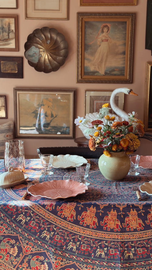 A styled dining table in a warm room setting, featuring a navy blue tablecloth with red and gold elephant motifs, pink plates, a floral centerpiece in a green vase, and a white swan figurine.