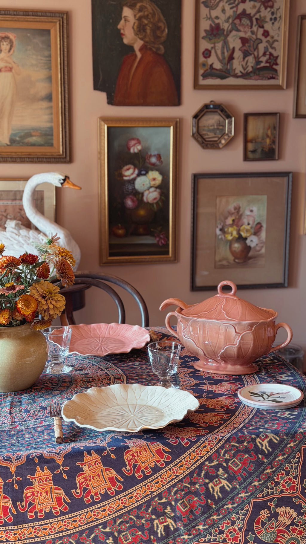 A styled dining table in a warm, vintage-inspired room setting, featuring a pink ceramic tureen, matching pink and cream plates, a patterned blue and orange tablecloth, and a white swan figurine against a wall covered in framed art.