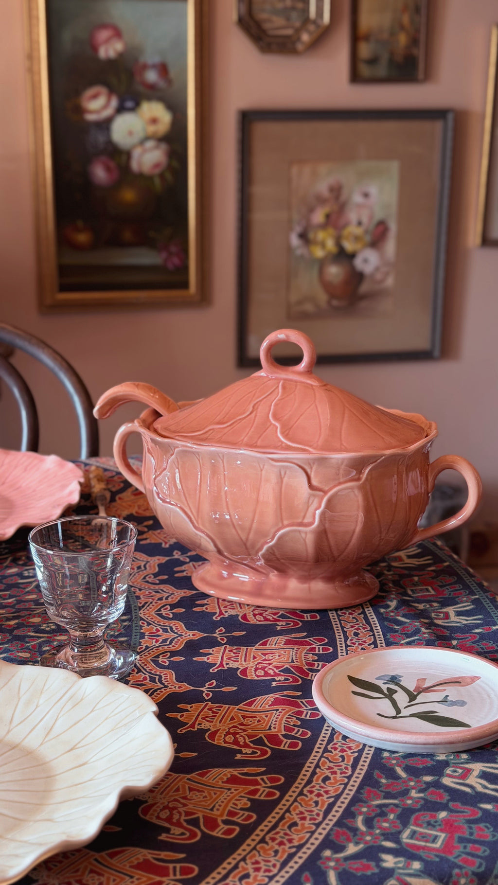 A pink ceramic lidded tureen with two handles sits on a patterned table runner in a dining room setting, surrounded by plates and a glass.