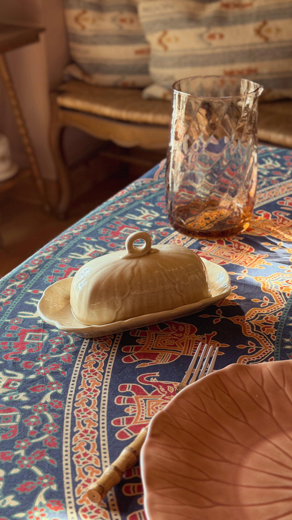 A Metlox Vernon Ware Lotus Sand Covered Butter Dish with a lid and small loop handle, set on a patterned blue and orange tablecloth next to a textured amber glass tumbler.