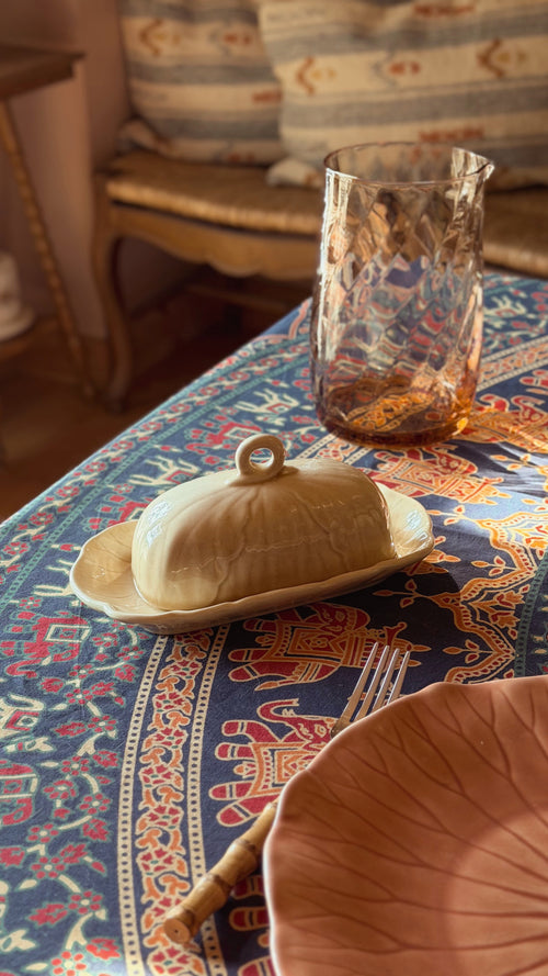 A Metlox Vernon Ware Lotus Sand Covered Butter Dish with a lid and small loop handle, set on a patterned blue and orange tablecloth next to a textured amber glass tumbler.