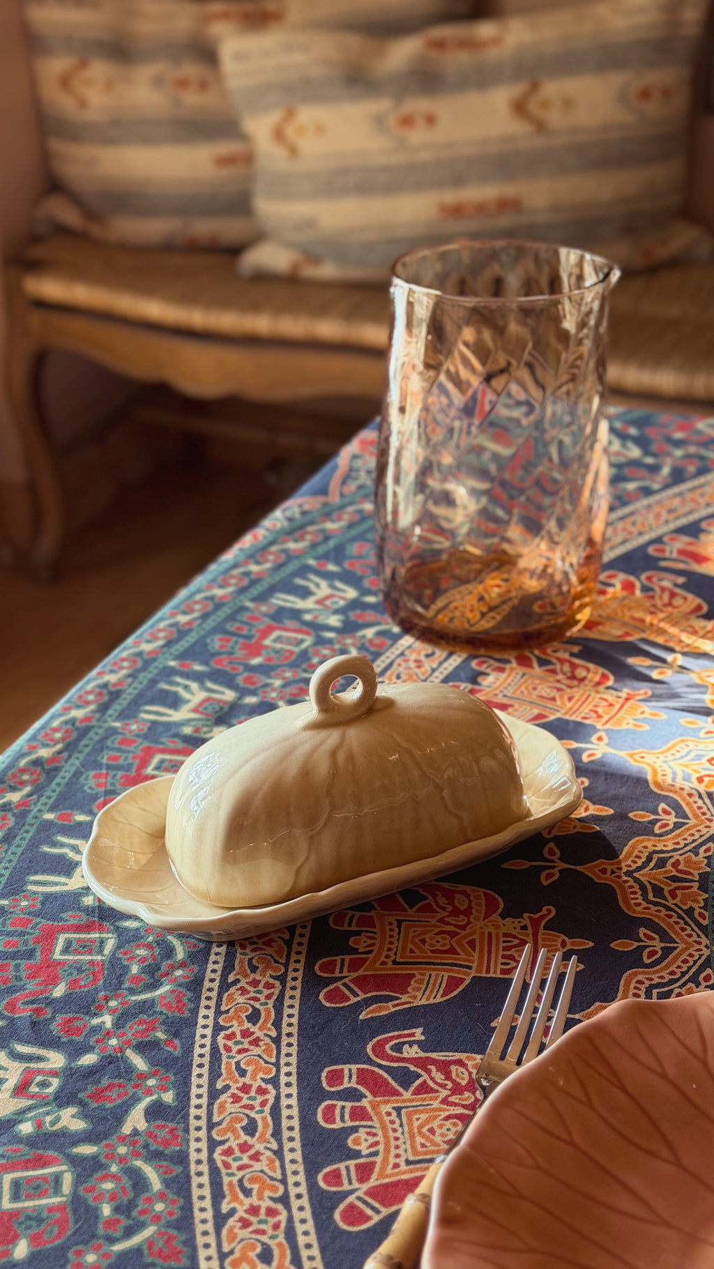 A Metlox Vernon Ware Lotus Sand Covered Butter Dish with a lid and loop handle, set on a patterned blue and red tablecloth next to a textured amber glass tumbler.