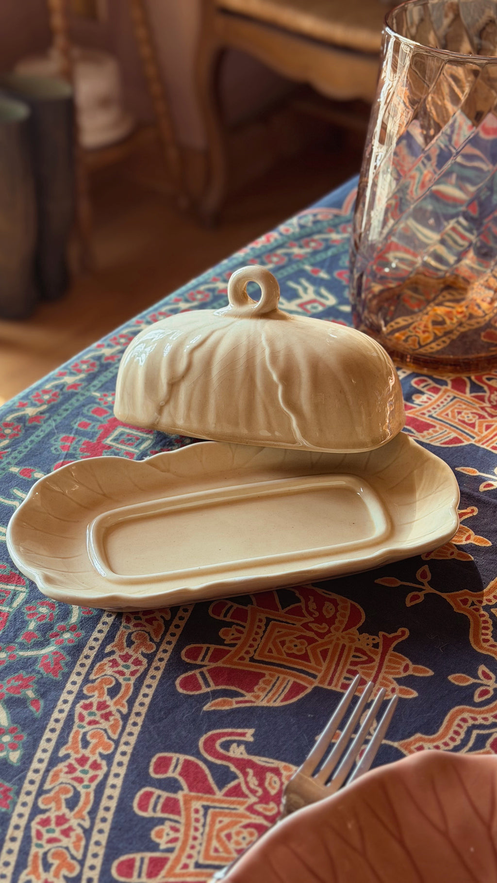 A ceramic butter dish with a matching lidded cover, shown on a patterned tablecloth in a dining setting. A fork is partially visible in the foreground, and a glass vessel is in the background.