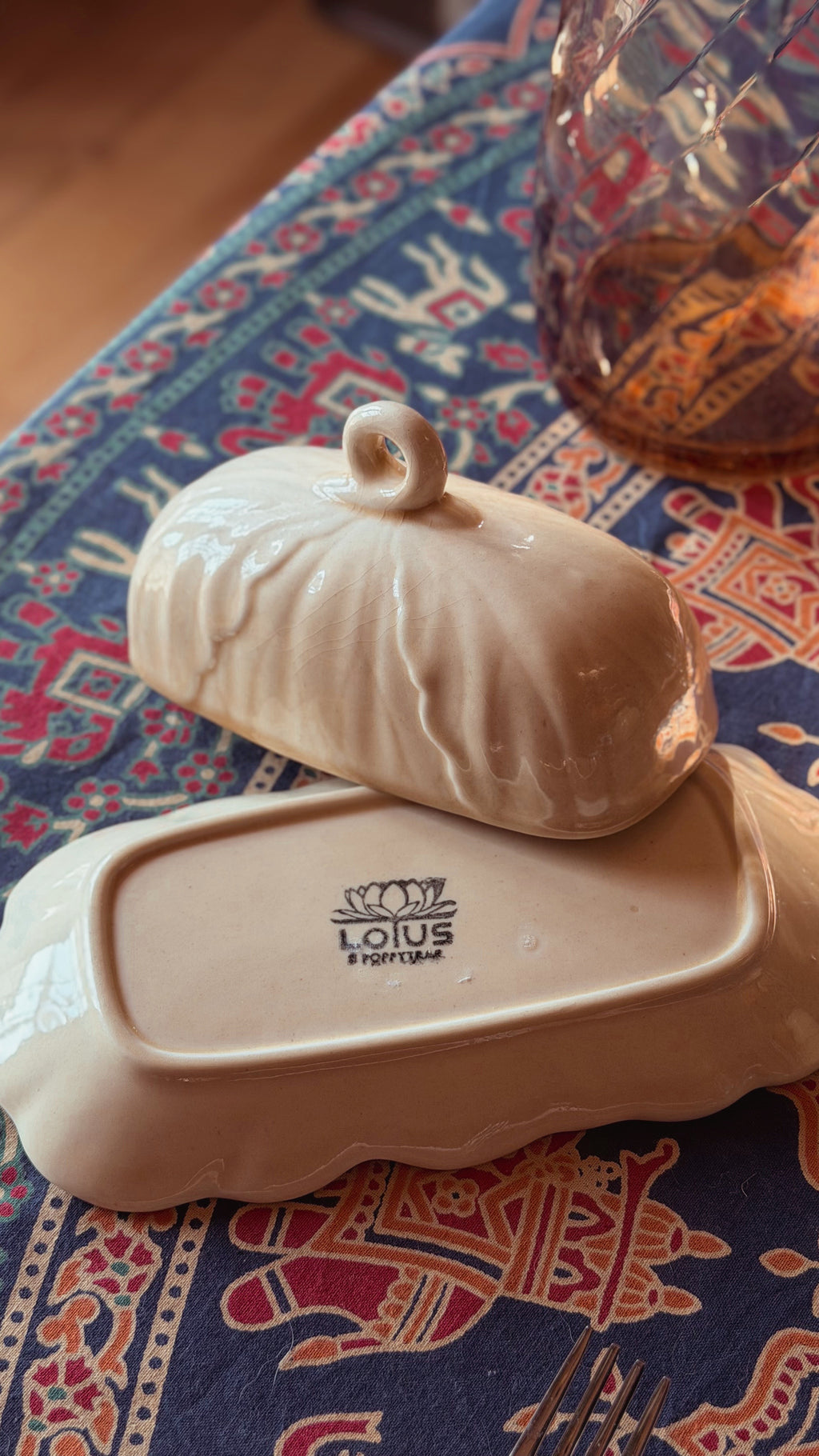 A cream-colored ceramic butter dish with a matching lidded cover, resting on a patterned blue tablecloth. The brand name 'LOTUS' is printed on the bottom of the dish.