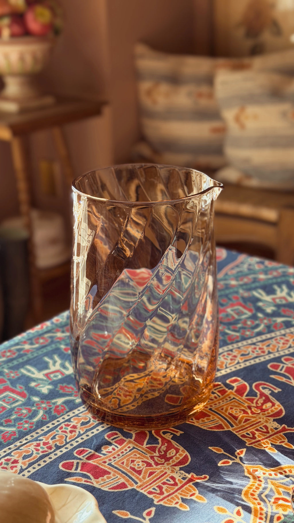 A Vintage Pink Swirl Optic Glass Pitcher with a twisted, textured design and a small pouring spout, filled with amber-colored liquid, set on a patterned blue tablecloth in a warm, cozy room.