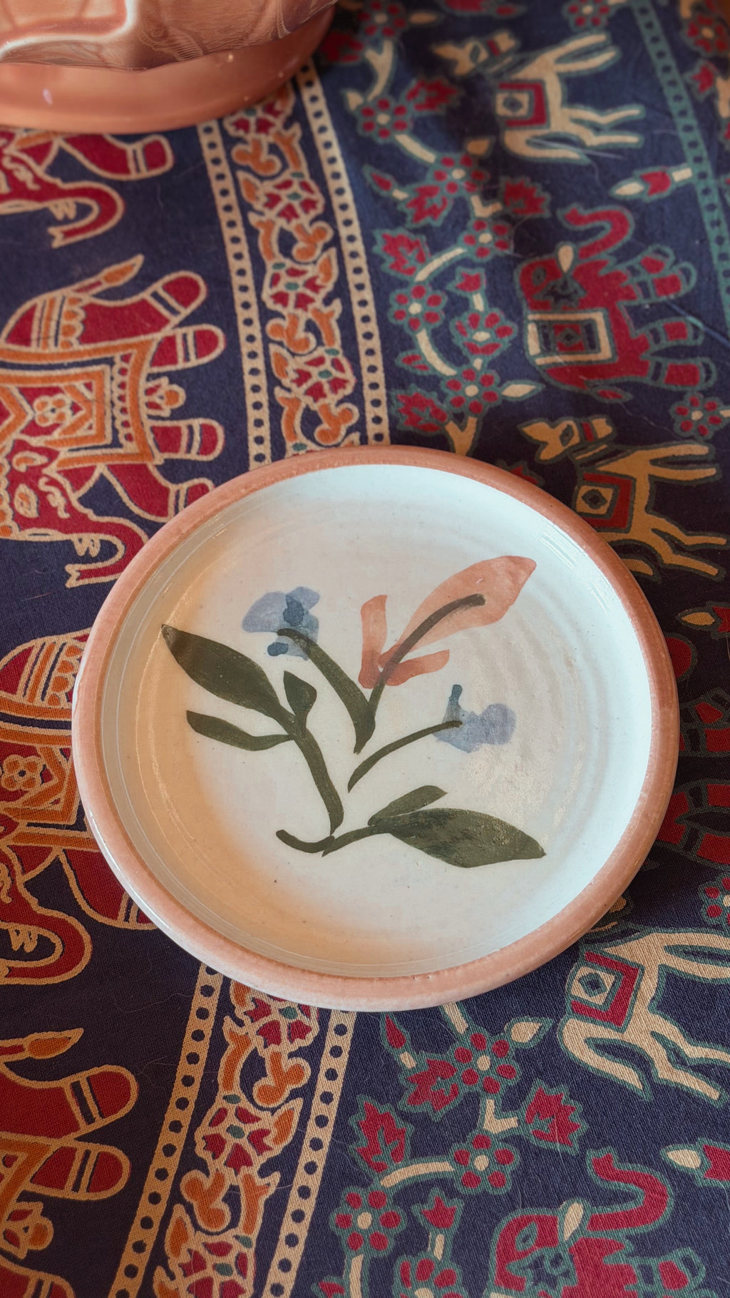 A round handmade ceramic plate with a natural terracotta rim and a hand-painted floral design in green, blue, and peach on a white glaze, resting on a patterned navy blue textile.