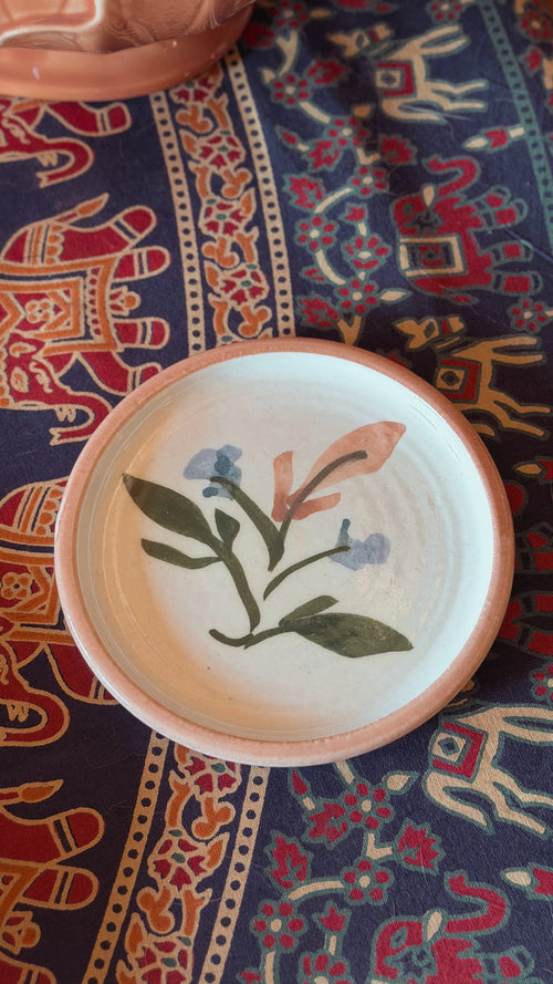 A round handmade ceramic plate with a natural terracotta rim and a hand-painted floral design in green, blue, and peach on a white glaze, resting on a patterned navy blue textile.