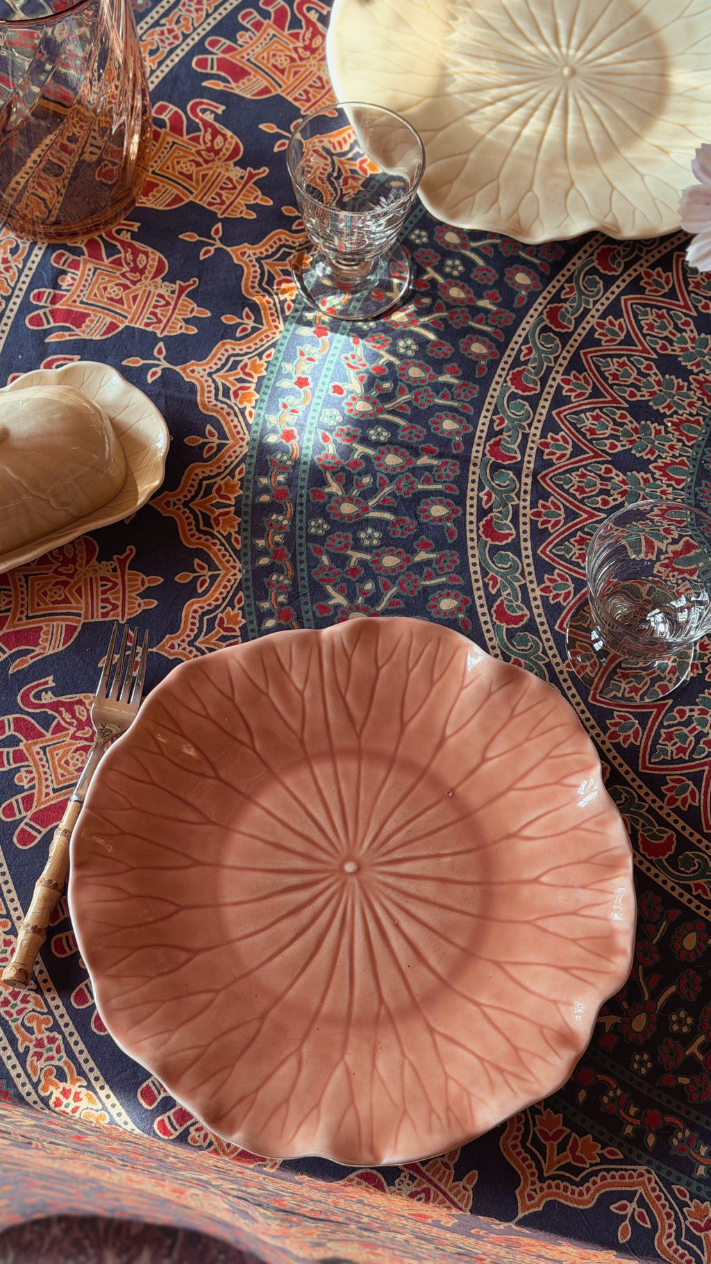 A terracotta-colored ceramic plate with a leaf-like radial pattern, styled on a dark patterned tablecloth alongside a glass, a fork, and other tableware.
