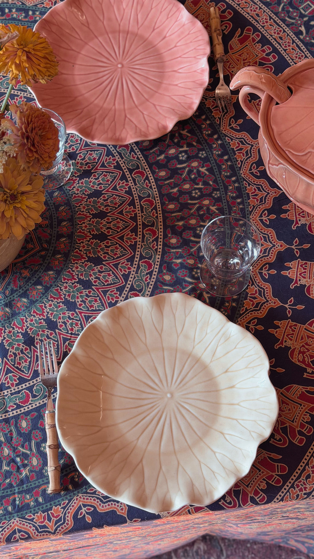 A styled tabletop arrangement featuring ceramic plates and a matching pink pitcher on a patterned blue and red tablecloth, with flowers and a glass in the scene.