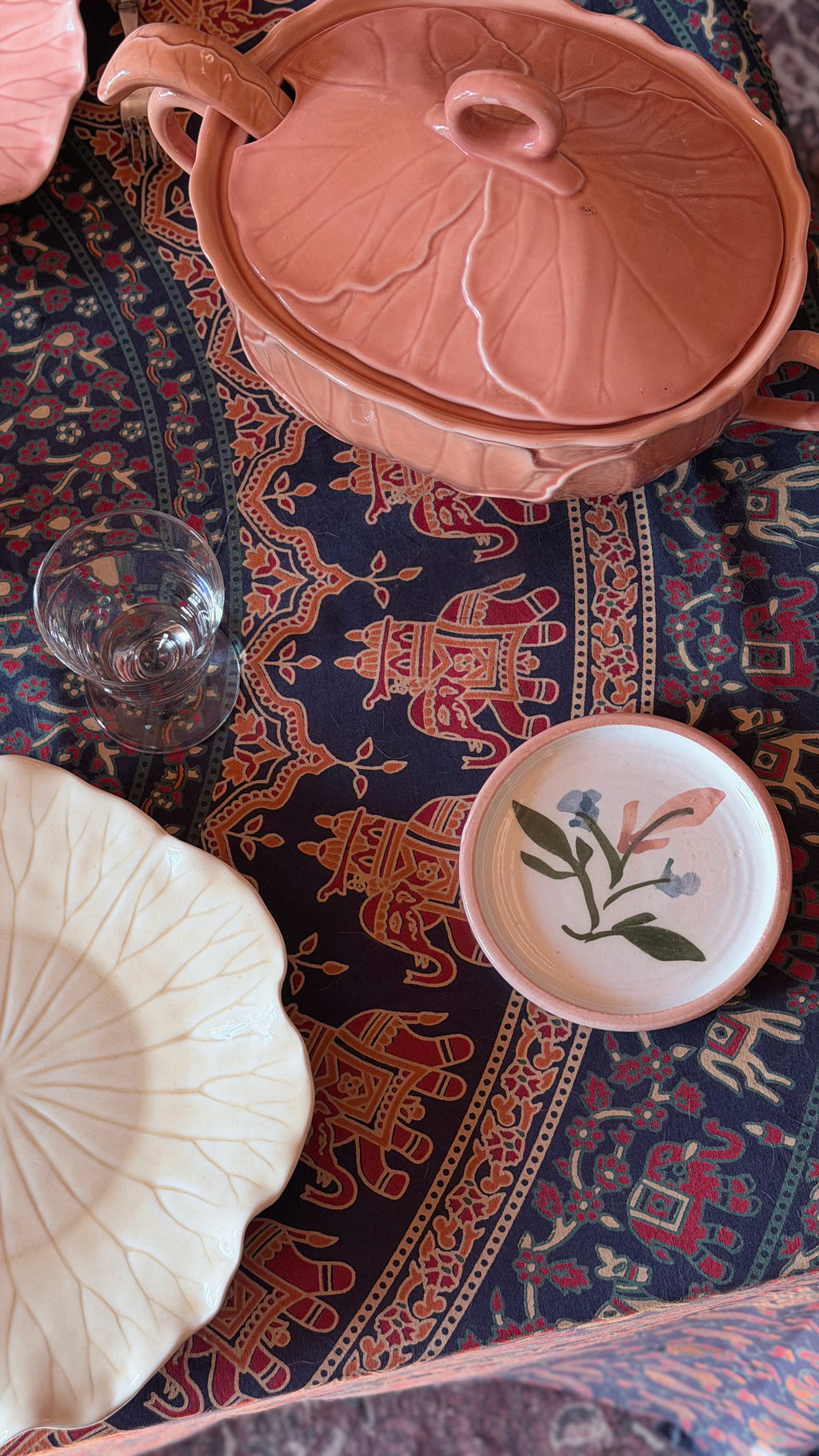 A styled flat-lay of ceramic tableware on a patterned rug, featuring a dusty pink leaf-shaped lidded pot, a cream-colored leaf-shaped plate, a small floral-patterned bowl, and a clear drinking glass.