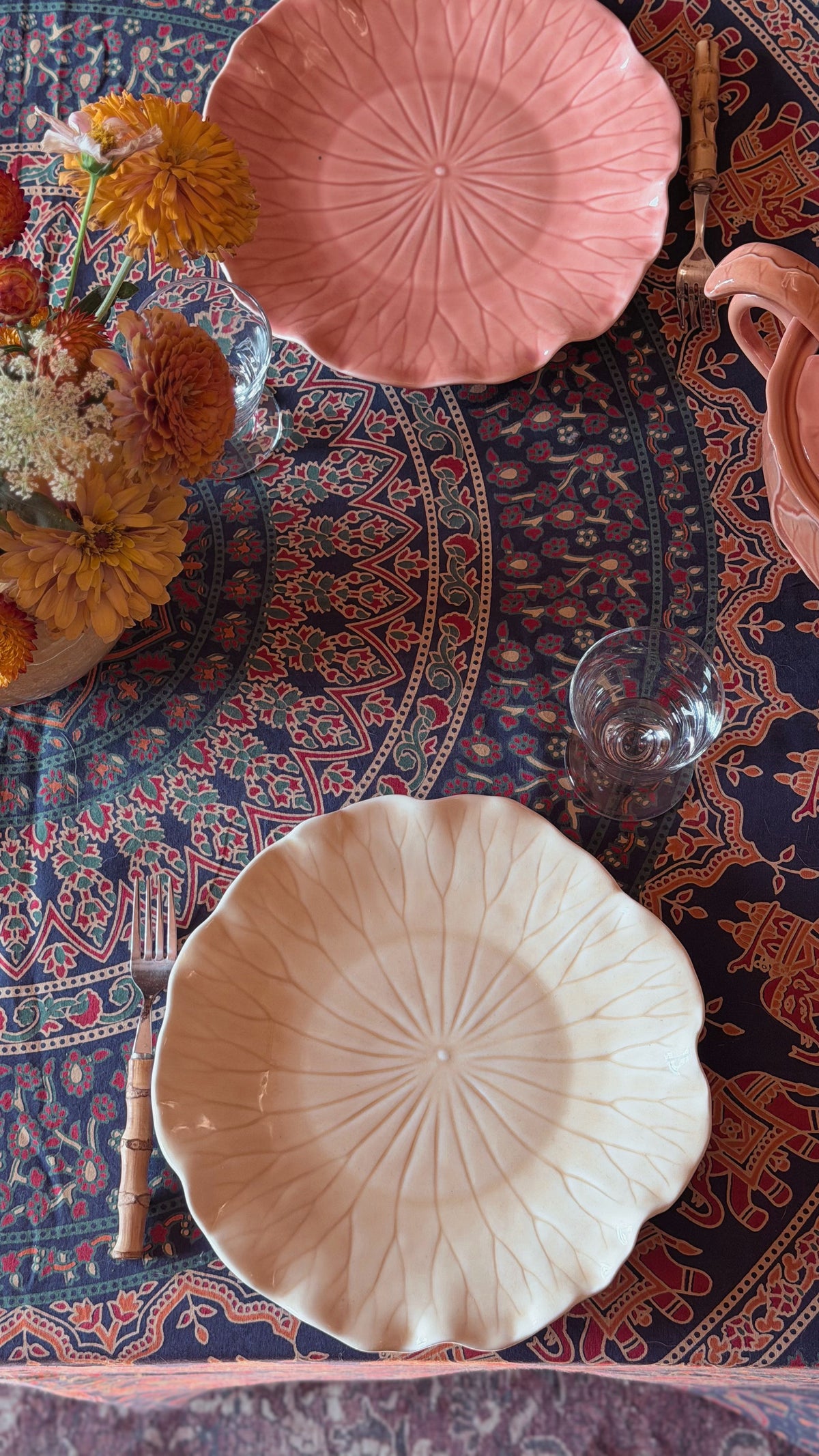 A top-down view of a styled table setting featuring two textured ceramic plates with a sunburst pattern, one in dusty pink and one in cream, arranged on a dark patterned tablecloth with flowers and glassware.