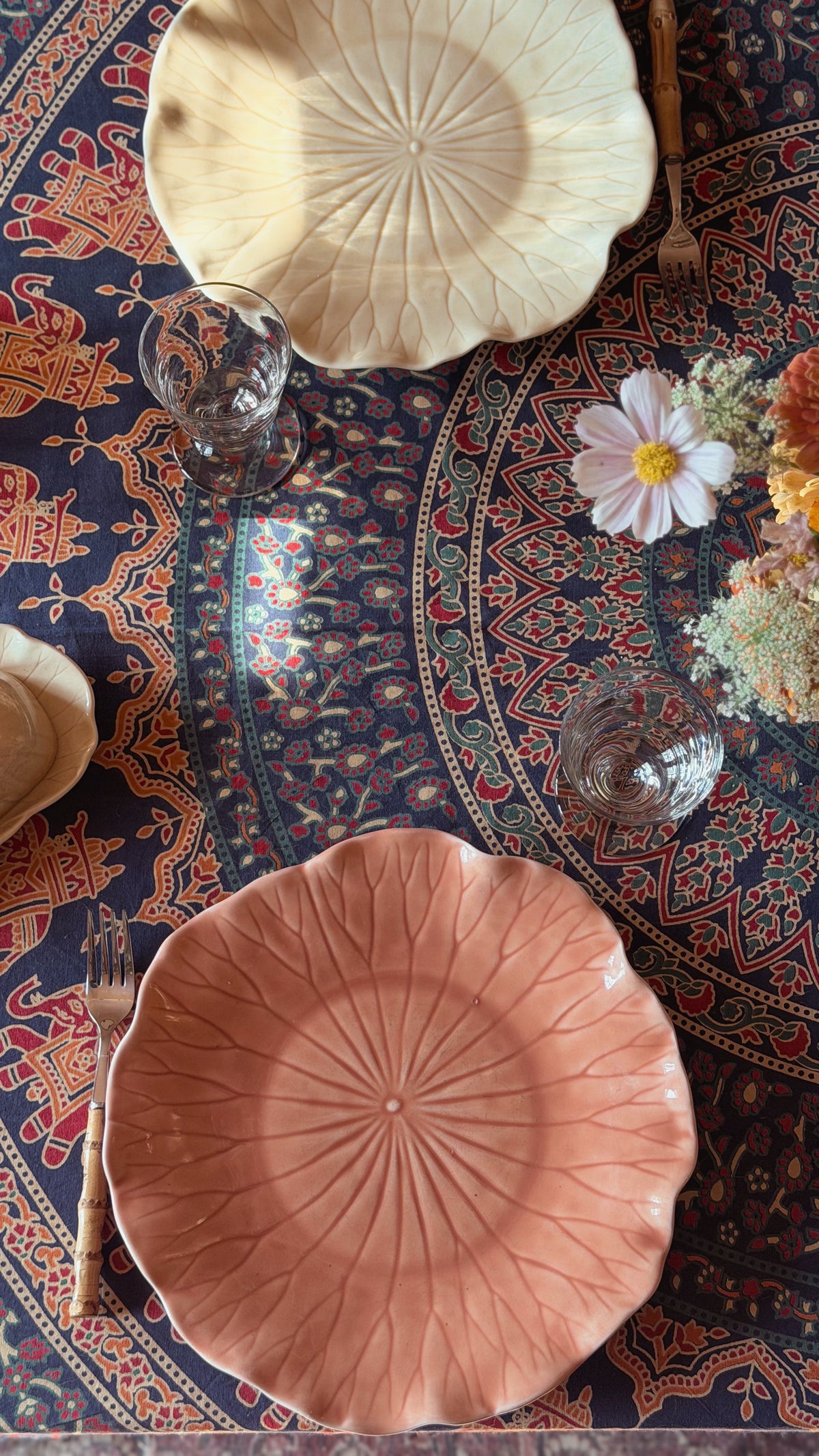 A styled overhead view of a table setting featuring two textured ceramic plates with a sunburst pattern, one in terracotta pink and the other in cream, placed on a dark blue patterned rug alongside glassware and flowers.