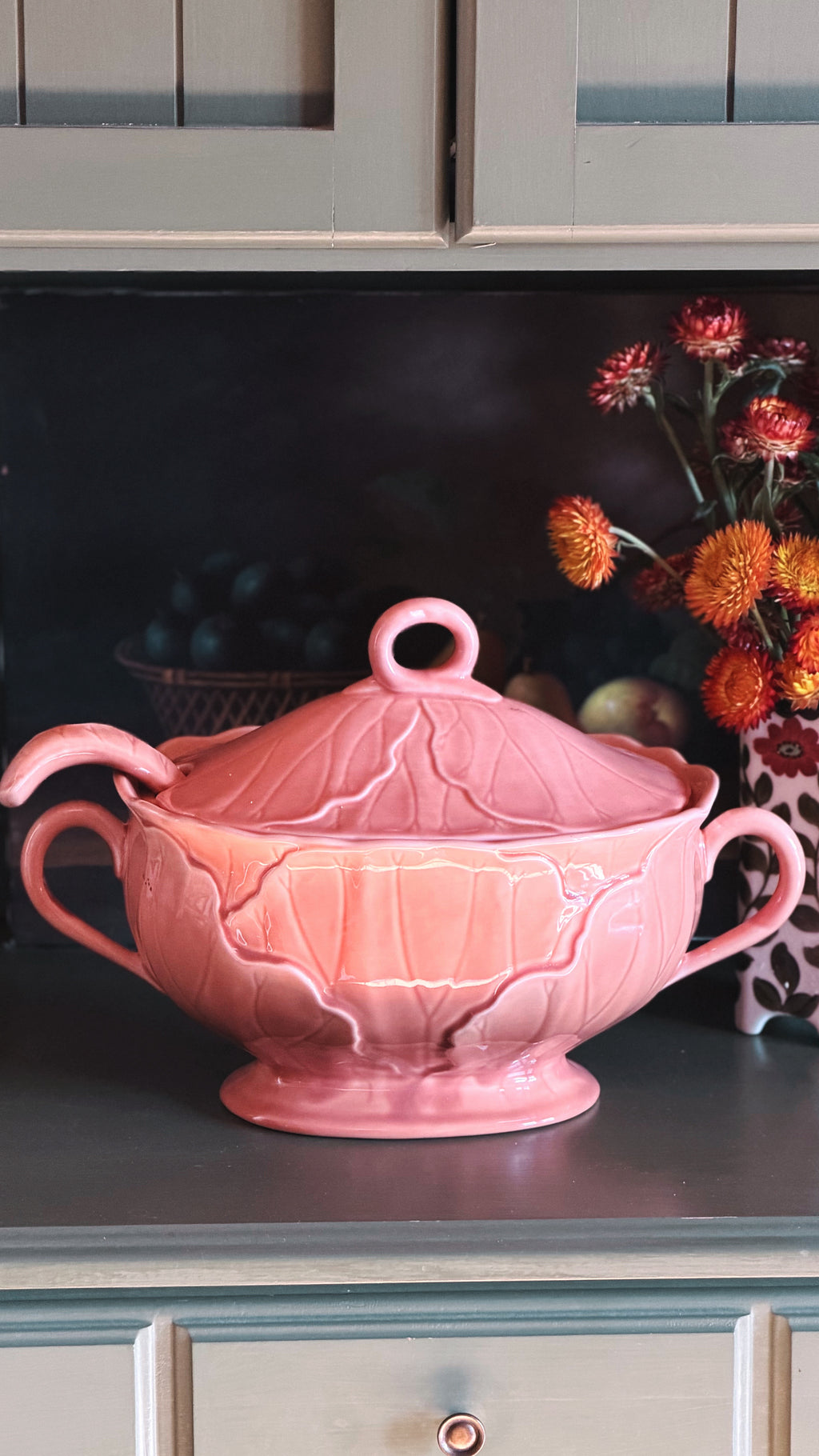A pink ceramic tureen with a matching lid, featuring embossed leaf-like detailing and two side handles, displayed on a dark countertop in a kitchen setting.