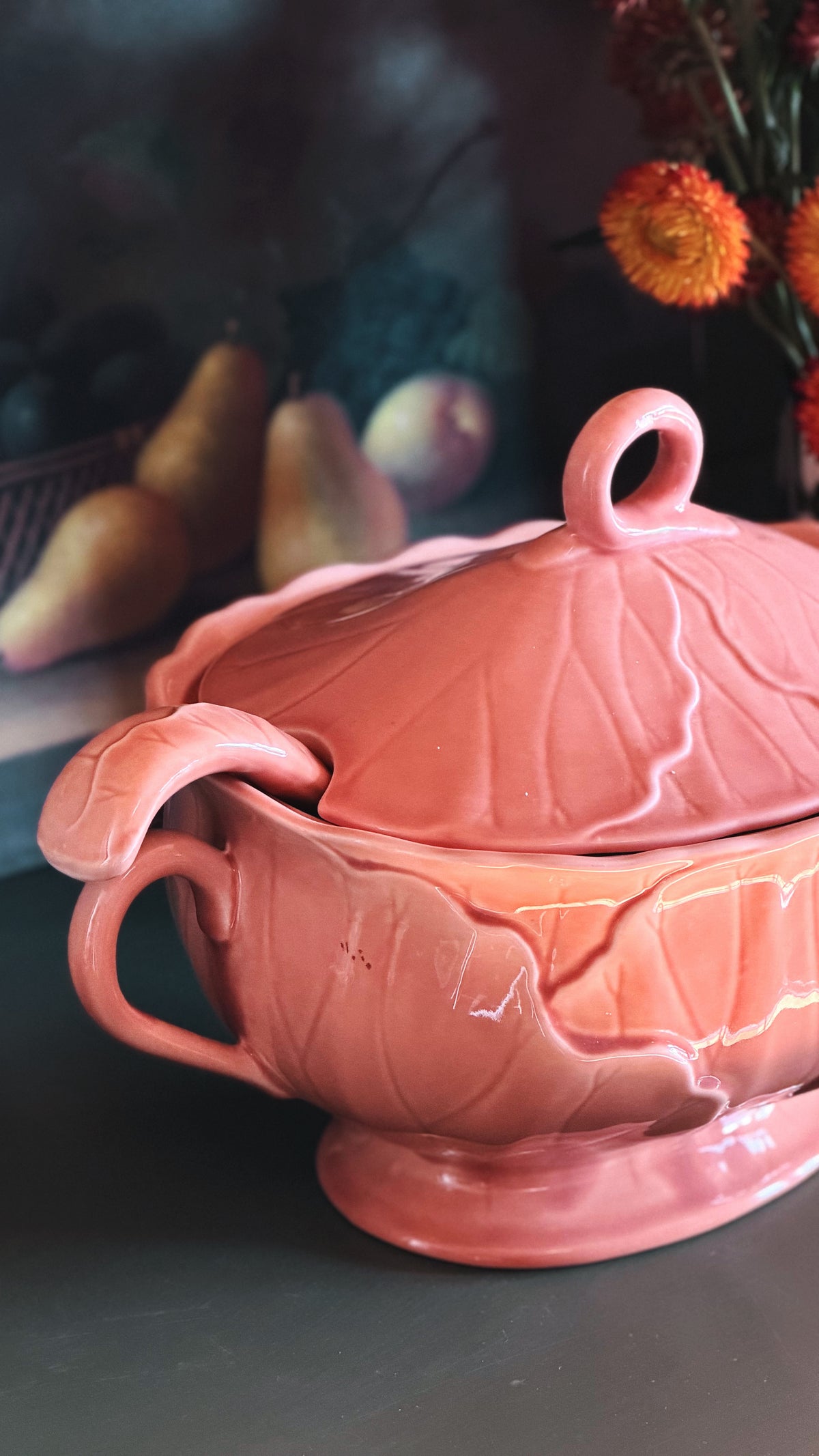 A glossy apricot pink ceramic tureen with a leaf-inspired design, featuring a lid with a loop handle, a side handle, and a spoon resting inside, set against a dark, softly blurred background with decorative elements.