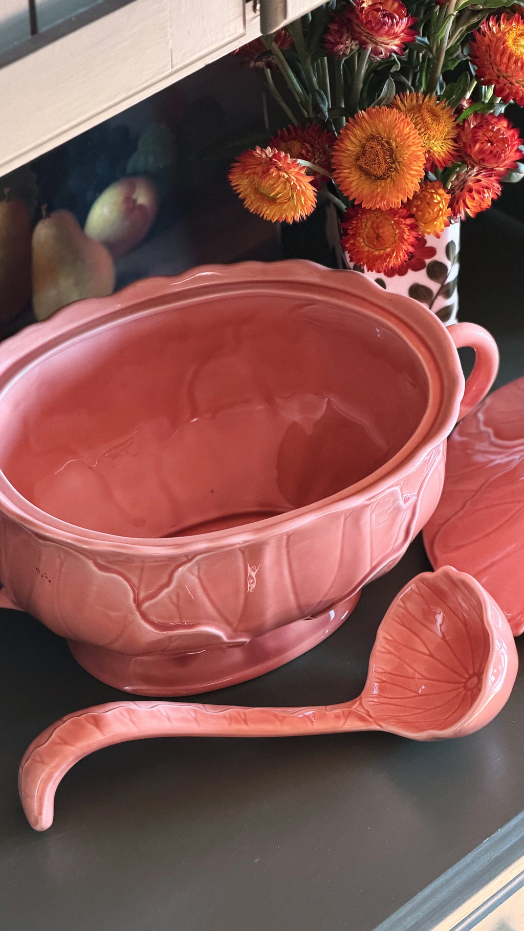 A pink ceramic serving bowl with a matching ladle and lid, displayed on a dark countertop with flowers in the background.