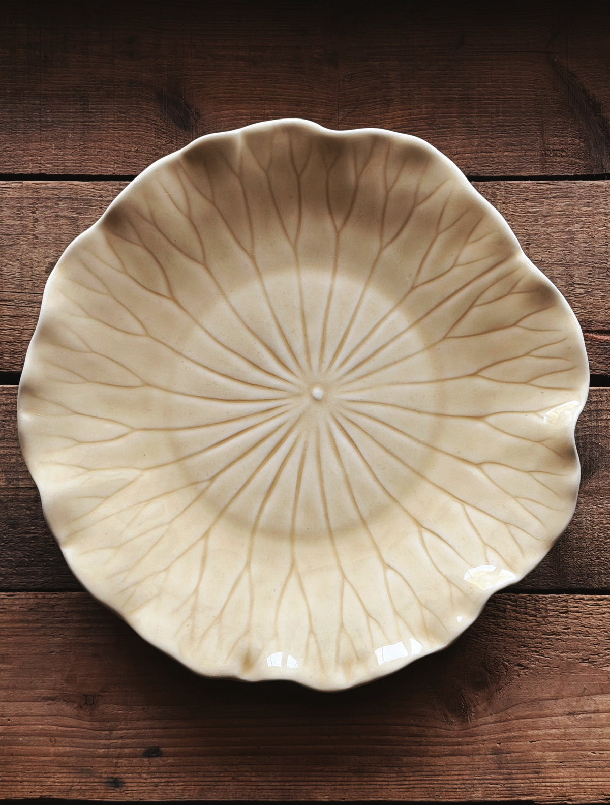 A Vintage Metlox Vernon Ware Lotus Sand Dinner Plate with a lotus leaf design, featuring a scalloped rim and radiating leaf vein patterns, shown from a top-down view on a dark wooden background.