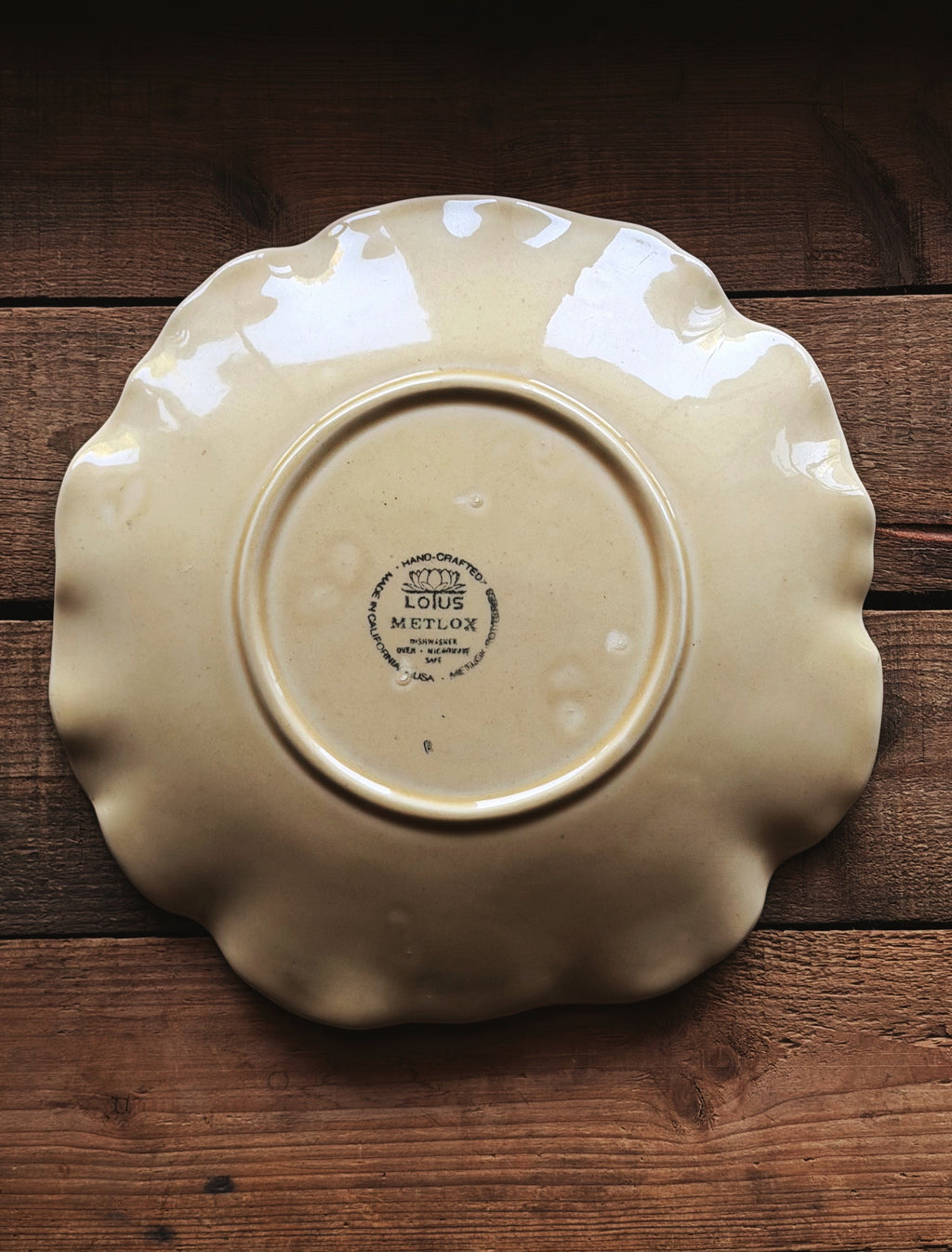 A Vintage Metlox Vernon Ware Lotus Sand Dinner Plate with a scalloped rim, photographed from above on a rustic dark wood background. The plate is cream-colored with a glossy finish and features a circular clay stamp in the center reading '1918 LOUIS MERLIAS'.