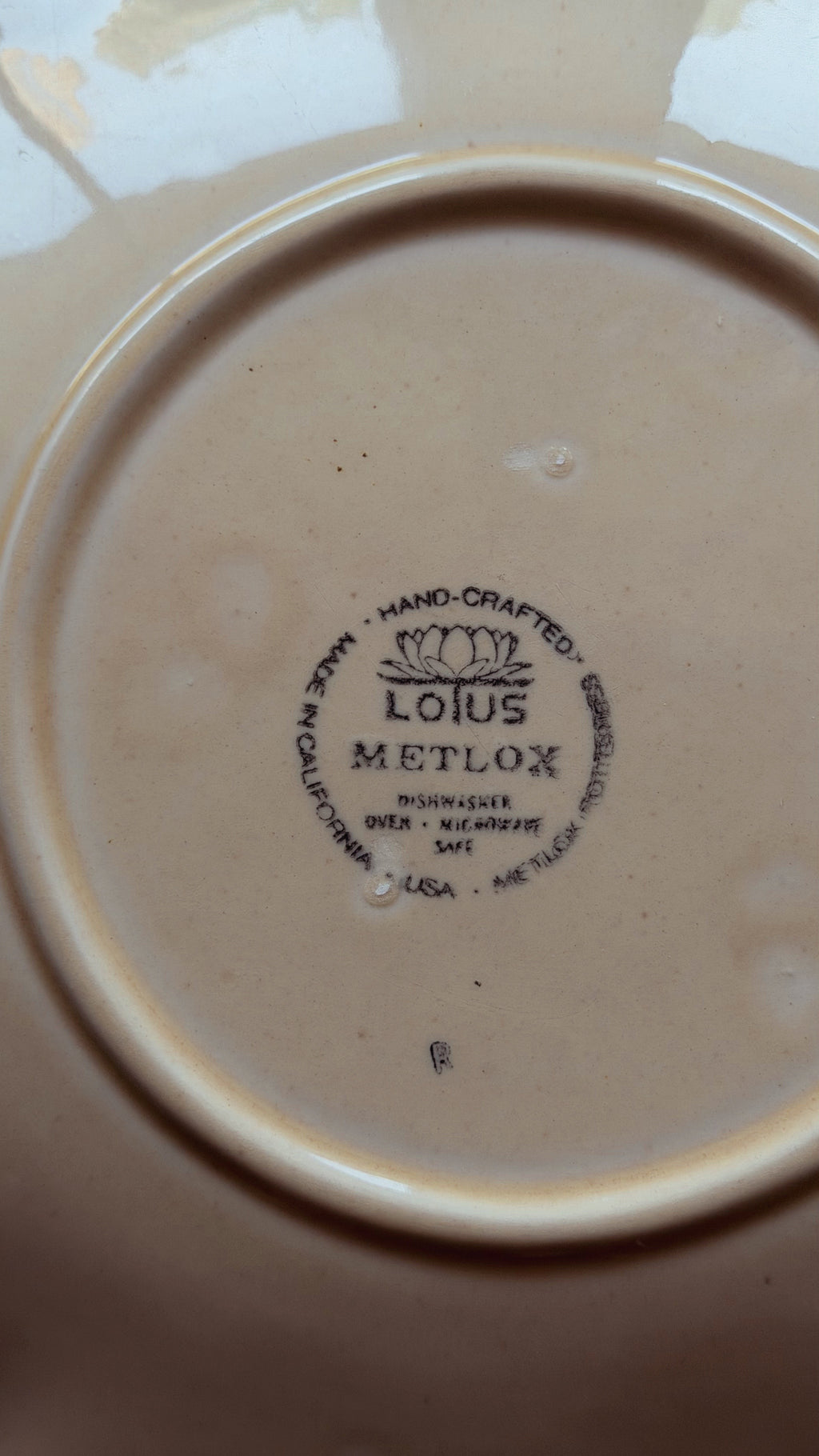 Close-up of the bottom of a beige ceramic plate or dish, showing the printed LOTUS METLOX brand mark in the center.