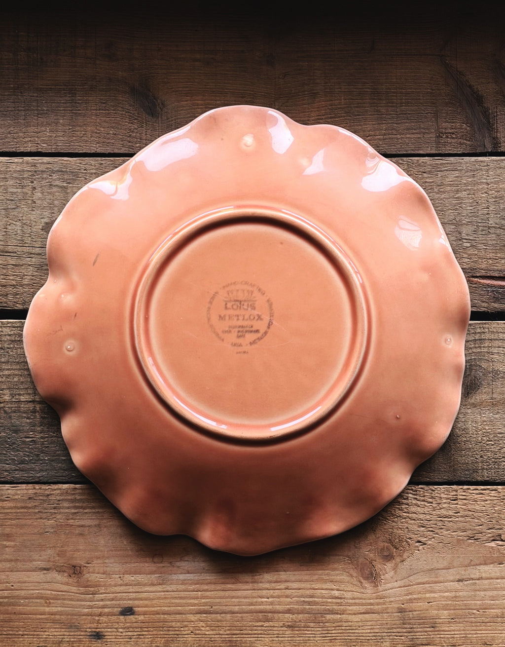 A scalloped ceramic plate in a warm terracotta or peach-orange glaze, photographed from directly above on a rustic dark wood plank surface. The center of the plate features a circular stamp reading 'LOTUS METLOX'.