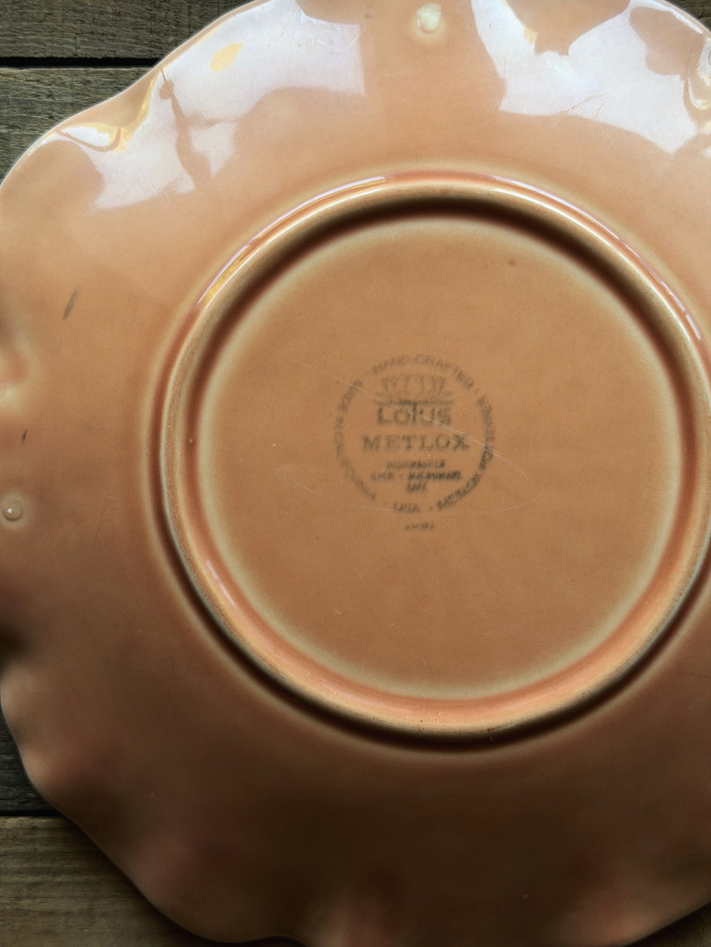 Close-up of the bottom of a vintage Lotus Metloxceramic plate or dish, showing a glossy terracotta pink glaze and the circular brand stamp in the center, resting on a dark wooden surface.